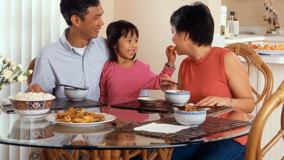 An Asian family, an adult male and female are seated around a table eating a meal with a young female standing in between the adults.